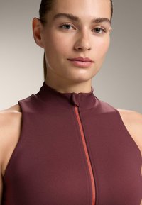 Bordó ujjatlan felső sima anyagból, magas gallérral és elöl narancssárga díszítésű cipzárral, közeli kép a nyakkivágásról és az arcról.