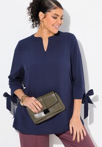 Blusa blu con maniche a tre quarti e lacci laterali, abbinata a una pochette verde oliva. Tessuto morbido, scollo a V e pantaloni a fantasia.