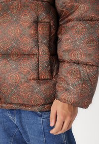 Gevoerde jas met een ingewikkeld multikleurig geometrisch patroon in roest, groen en rood. Heeft een boxy vorm en een elastische zoom.