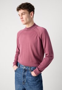 Pulover dolgih rokavov, oprijet kroj v mauve pletenini z visokim ovratnikom. Ima raglan rokave in rebraste manšete. V kombinaciji z modrimi jeans hlačami.