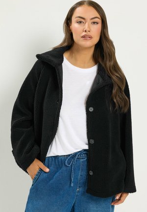 Vrouw met lang bruin haar die een zwarte fleecejas draagt over een wit overhemd en blauwe jeans met trekkoord, staand tegen een effen achtergrond.