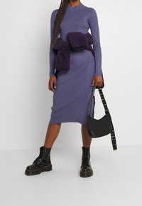 Abito a maglia ribattuta viola con maniche lunghe e colletto alto. Un spesso maglione viola scuro è legato in vita. Stivali neri con lacci e una borsa nera.