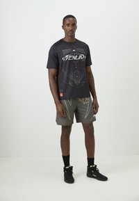 Camiseta deportiva negra con gráfico geométrico blanco, combinada con shorts grises con acentos claros y un ajuste holgado, y calzado negro.