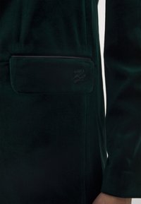 Grön sammet blazer med en lapplåda med diskret branding och en elegant, skräddarsydd design. Tyget har en mjuk, plyschliknande textur.