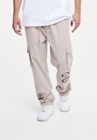 Beige cargobroeken met zijzakken, geborduurde patches, een trekkoord in de taille, gedragen met witte sneakers. Soepele, lichte stof.