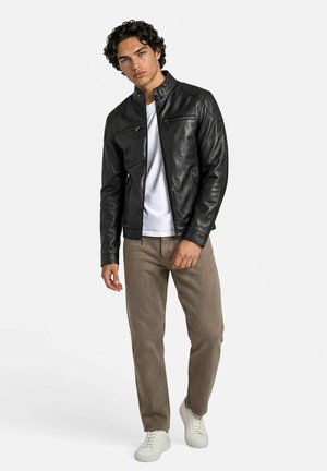 Jonge man in een zwarte leren jas, wit T-shirt, beige broek en witte sneakers, staand tegen een effen lichte achtergrond.