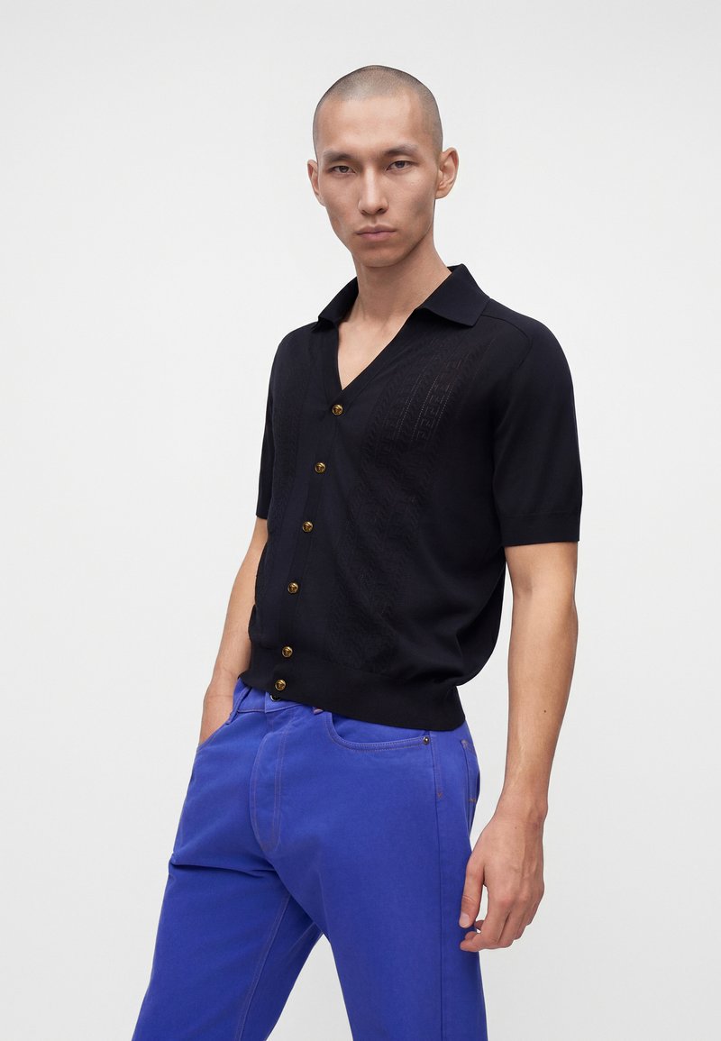 Homme à la tête rasée portant une chemise noire à manches courtes boutonnée et un pantalon bleu vif, debout avec une main dans la poche.