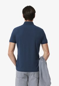 Mörkblå polo med korta ärmar, slät textur, krage och rak nederkant. Bäraren håller en grå tröja över armen.