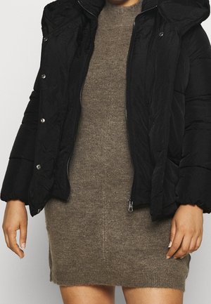 Czarna kurtka puchowa z kapturem na dopasowanej, sięgającej kolan brązowej dzianinowej sukience, noszona przez osobę na neutralnym tle.