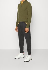 Mies, joka wearing oliivinvihreä varttinveto neule, keltainen paita, mustat kapeat housut, valkoiset urheilukengät, seisoo vaaleaa seinää vasten.