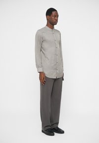 Άνδρας στέκεται φορώντας ένα μοτίβο πουκάμισο με μακριά μανίκια, χαλαρές γκρι παντελόνες και μαύρα αθλητικά παπούτσια, μπροστά σε λευκό φόντο.