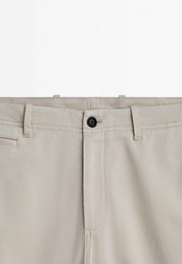 Section de taille d'un pantalon beige avec une fermeture à bouton noir, des passants de ceinture et une petite poche horizontale sur le côté gauche.