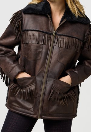 Bruine leren jas met zwarte schapenvachtkraag en franjes op de borst, mouwen en zakken, gedragen met de rits dicht en handen in de zakken.