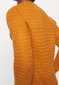 Gros plan sur une personne portant un pull en tricot orange texturé, montrant des motifs détaillés en torsade et en vague sur le dos et les manches.