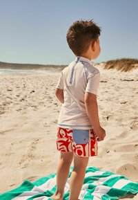 Barnens rash guard i ljust grått med en blå dragkedja, tillsammans med röda shorts med vita abstrakta mönster. Står på en strandhandduk.