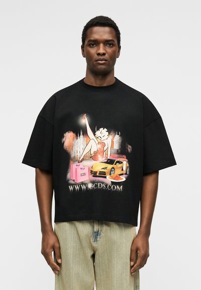 Άνδρας που φοράει μαύρο υπερμεγέθη μπλουζάκι με γραφικό μιας καρτούν γυναίκας, κίτρινο σπορ αυτοκίνητο, ροζ σακούλες αγορών και διεύθυνση URL ιστοσελίδας στο μπροστινό μέρος.