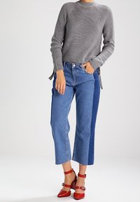 Grijze geribde trui, lichtblauwe en donkerblauwe twee-kleurige cropped jeans, en rode enkelschoenen met bandjes en een vierkante neus.