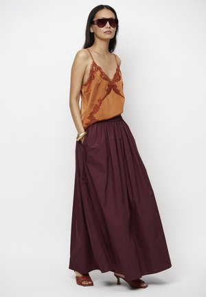 Vrouw met grote zonnebril, roestkleurige satijnen top met kantdetails, lange bordeauxrode rok, gouden armband en sandalen met hak.