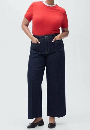 Personne portant une chemise rouge à manches courtes rentrée dans un jean taille haute à jambes larges bleu foncé avec poches, et des chaussures plates noires sur fond gris.