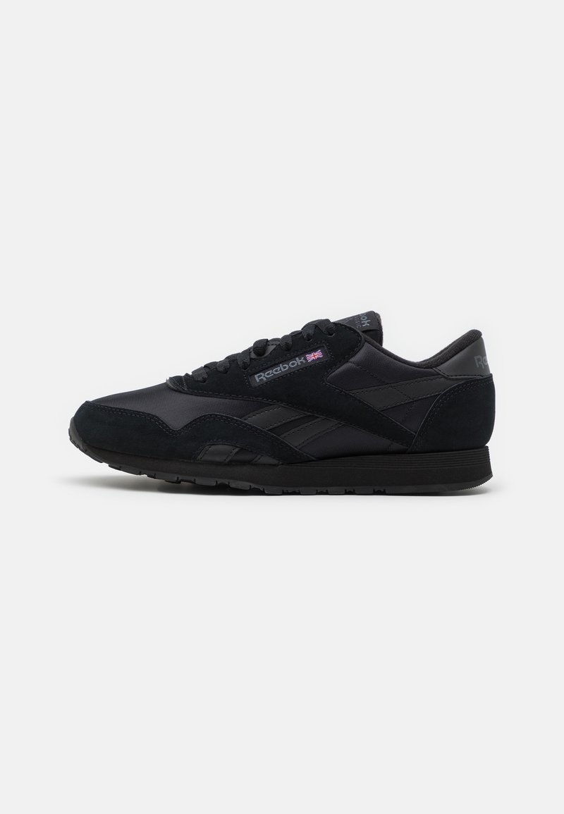 Baskets noires Reebok avec lacets à l’avant, logo et drapeau britannique sur le côté, semelle texturée et col rembourré sur fond blanc.