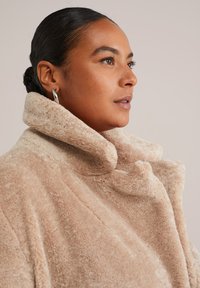 Manteau en fausse fourrure beige avec un col haut et une texture douce. Présente une coupe décontractée et des coutures subtiles, mettant en avant la chaleur et le confort.
