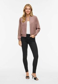 Femeie cu păr lung blond poartă o jachetă bomber cu fermoar de culoare mov, top alb, blugi skinny negri și sandale negre cu toc, stând în fața unui fundal alb.