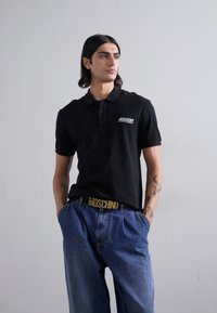 Čierna polokošeľa s trojradovým gombíkovým zapínaním a bielym logom "MOSCHINO". Nosená s modrými džínsami s vysokým pásom a zlatým opaskom.