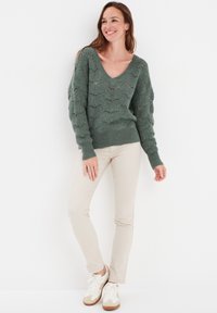 Un pull en tricot vert avec un décolleté en V profond, un motif de vagues texturé et des manches amples, associé à un pantalon beige clair et des baskets blanches.