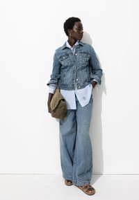 Veste en denim, bleu clair, avec deux poches poitrine. Superposée à une chemise à rayures bleues. Jean bleu clair à coupe décontractée. Sac à épaule vert olive.