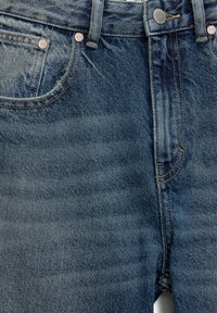 Gros plan sur un jean en denim bleu montrant les poches avant, les passants de ceinture et les rivets métalliques avec des coutures apparentes et une légère décoloration du tissu.