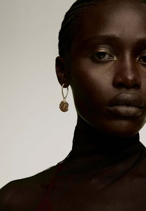 Pendientes de aro de oro con un colgante circular con un diseño en relieve; modelo vistiendo una blusa negra transparente. El fondo es neutro.
