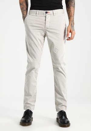 Mies pukeutunut vaaleanbeigeihin chinoihin, mustiin nahkakenkiin ja mustaan paitaan, seisoo osittain taskut kädessään.