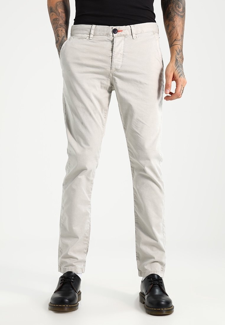 Un homme portant un pantalon chino beige clair, des chaussures en cuir noir et une chemise noire, debout les mains partiellement dans les poches.