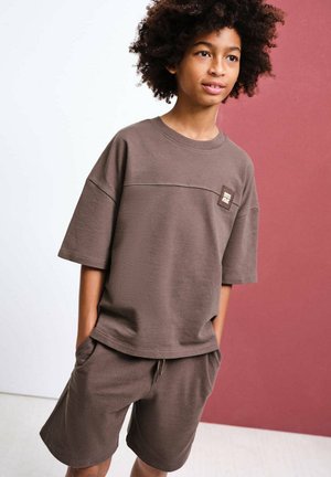 Junge mit lockigem Haar, der ein locker sitzendes braunes T-Shirt und passende Shorts trägt, steht mit den Händen in den Taschen vor einem weißen und kastanienbraunen Hintergrund.