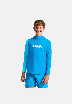 Teenager-Junge lächelt, trägt ein hellblaues Langarmshirt mit weißem "SLAM"-Logo und passende blaue Shorts vor einem einfarbigen Hintergrund.