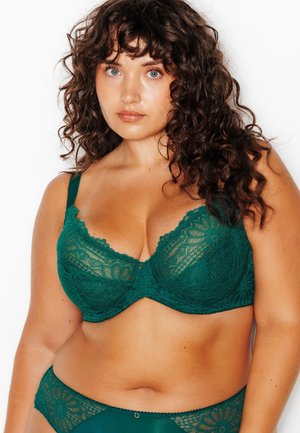 Donna con capelli castani ricci che indossa un reggiseno di pizzo verde e biancheria intima abbinata su uno sfondo bianco semplice.