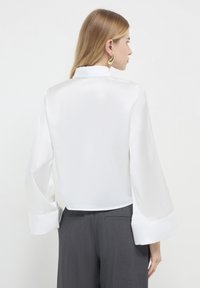 Blouse en satin blanc avec de longues manches larges et un col haut, associée à un pantalon gris ; texture lisse, design minimal, sans motifs visibles.