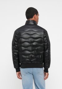 Giubbotto piumino nero con finitura lucida, caratterizzato da texture trapuntata, colletto alto e polsini a coste, indossato sopra jeans azzurri chiari.