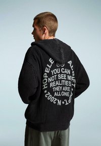Fekete kötött kapucnis pulóver, amelynek hátulján nagy fehér szöveg található, mondatokkal és kör alakú dizájnnal. Texturált, laza fazon.