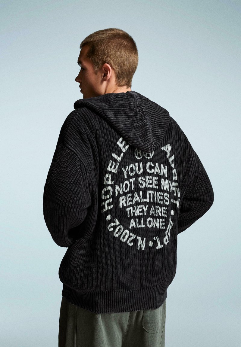 Fekete kötött kapucnis pulóver, amelynek hátulján nagy fehér szöveg található, mondatokkal és kör alakú dizájnnal. Texturált, laza fazon.