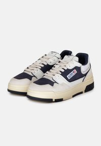 Baskets en daim blanches et bleu marine avec des accents beiges, lacets plats et semelles en caoutchouc noires. Présentent un patch logo sur le côté.