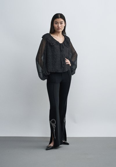 Γυναίκα σε όρθια στάση, φοράει ένα διάφανο μαύρο μπλουζάκι με βολάν και μαύρο παντελόνι με κεντημένα ανοίγματα κοντά στους αστραγάλους και μαύρα μυτερά παπούτσια.