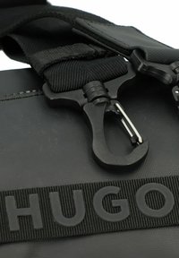 Borsa nera con superficie testurizzata. Presenta una tracolla con clip e la parola "HUGO" in rilievo in lettere maiuscole su una banda tessuta.