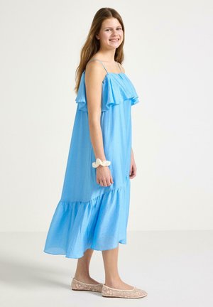 Smilende ung kvinde med langt hår iført en lyseblå ærmeløs kjole og beige balletsko, stående foran en ensfarvet hvid baggrund.