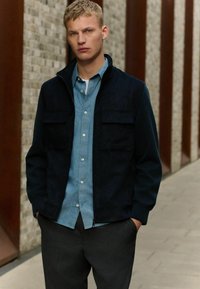 Chaqueta azul marino oscura con dos bolsillos en el pecho, superpuesta a una camisa de botones azul claro. El modelo lleva pantalones gris ajustados y está de pie frente a una pared texturizada.