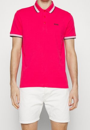 Homme portant un polo rose vif avec un col et des manches rayés, associé à un short blanc, debout devant un fond uni.