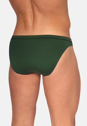 Mann, der dunkelgrüne Badehose trägt, gezeigt vom unteren Rücken bis zur Mitte des Oberschenkels vor einem einfarbigen Hintergrund.