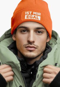 Orange knit beanie with white text "IST MIR EGAL," worn with a green puffer jacket and black inner layer, showcasing color contrast and texture.