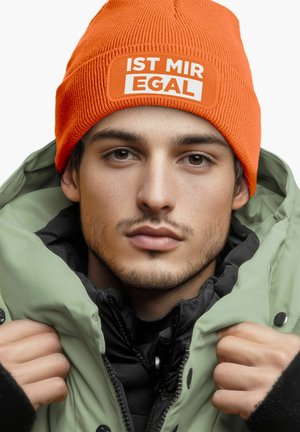 Orange knit beanie with white text "IST MIR EGAL," worn with a green puffer jacket and black inner layer, showcasing color contrast and texture.