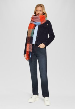 Veste en corduroy bleu foncé avec deux poches, chemise bleu clair, écharpe à carreaux multicolore, jeans foncés et baskets blanches avec semelle texturée.